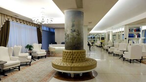 A lobby with a central column, white sofas, a patterned rug, and a bookshelf in the background. - Hotel Roxy Plaza (Soave)