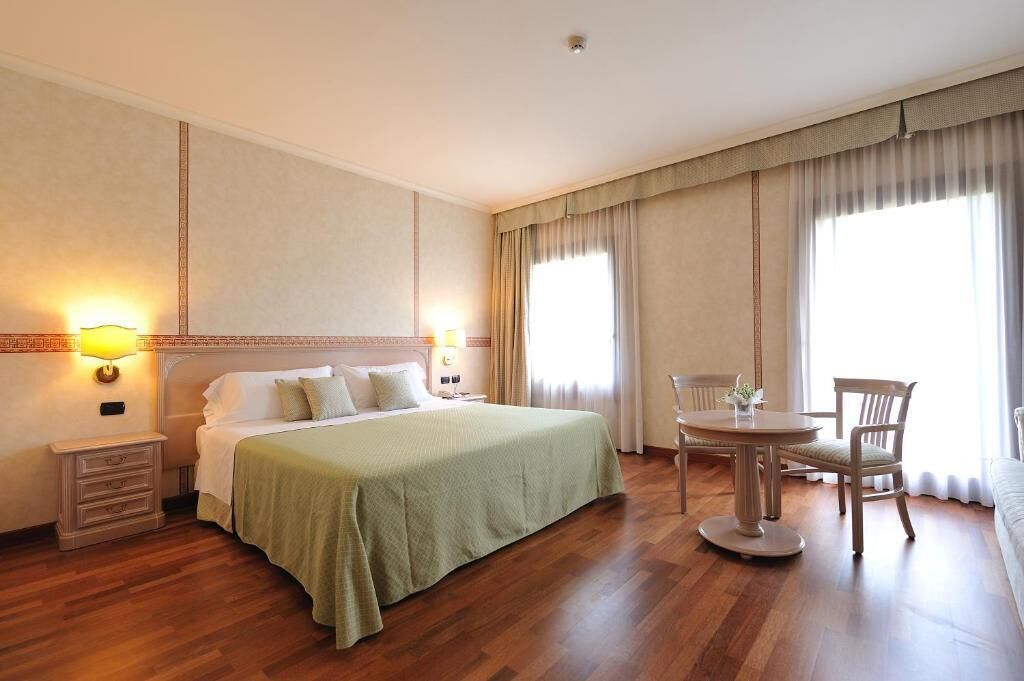 Quarto de hotel com uma cama, mesas de cabeceira, uma mesa de jantar redonda com cadeiras e janelas amplas com cortinas.