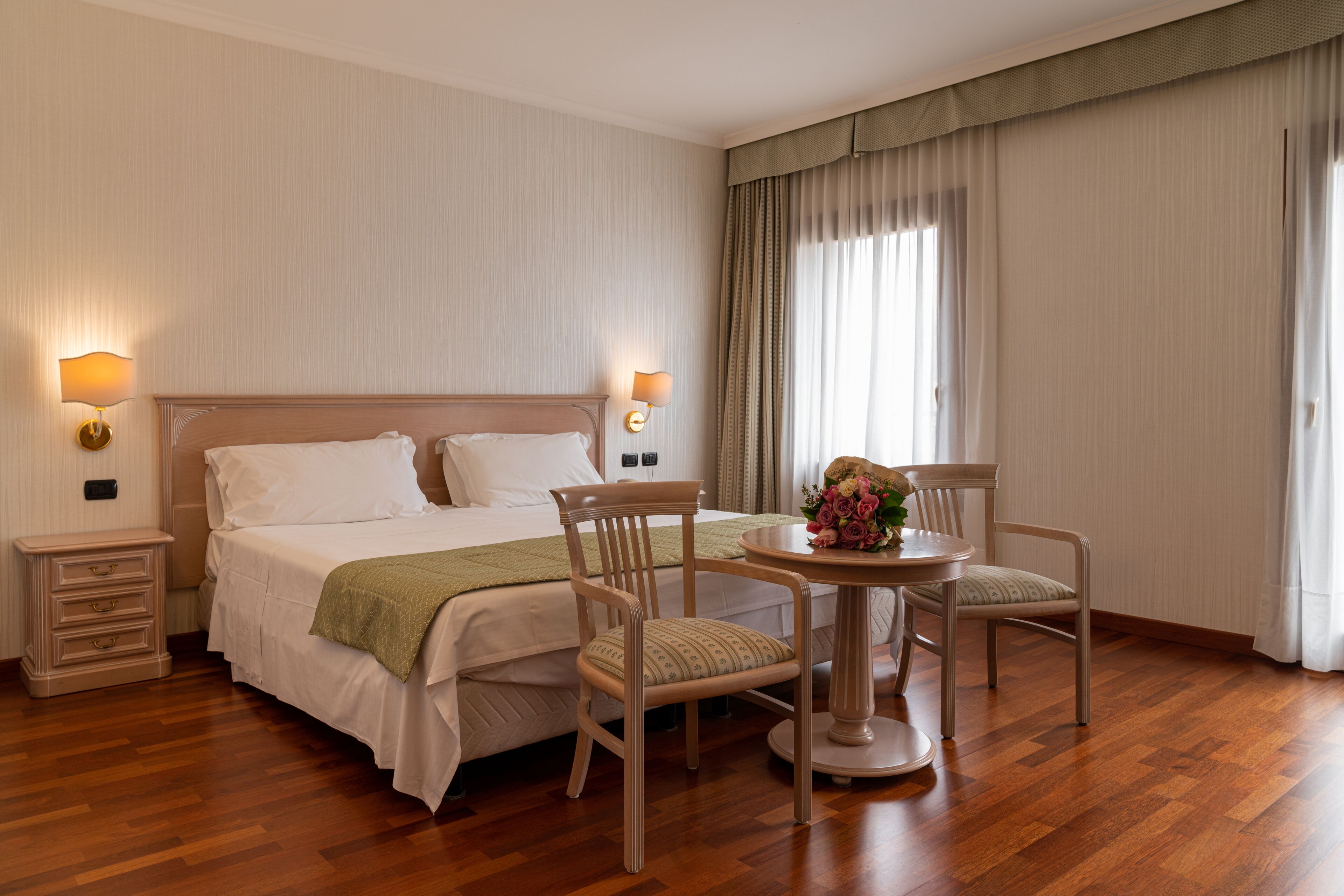 Habitación de hotel con una cama grande, dos mesitas de noche con lámparas, una mesa redonda con sillas y un ventanal con cortinas.