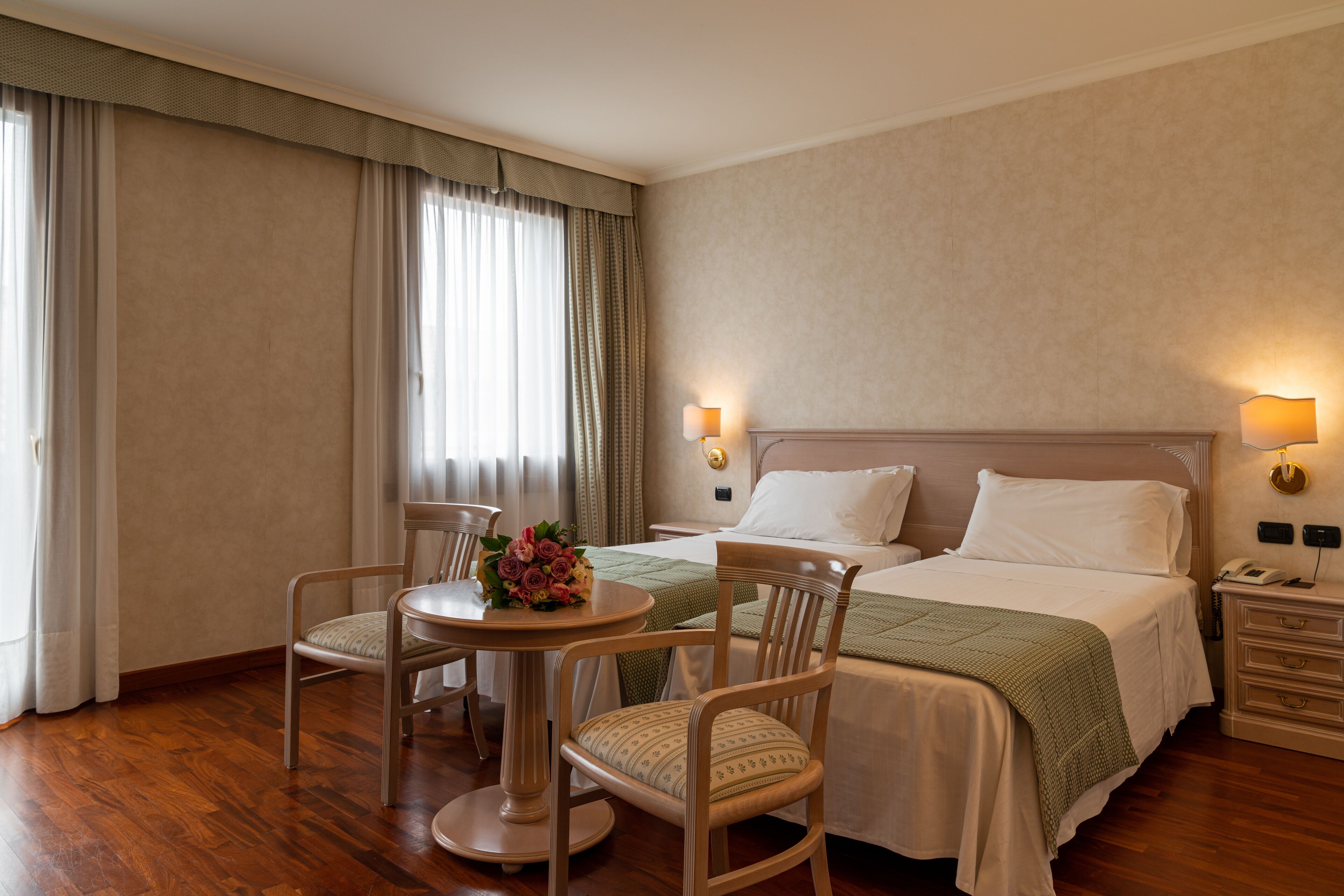 Ein Hotelzimmer mit einem Bett, zwei Stühlen, einem kleinen Tisch mit Blumenvase und einem Nachttisch mit Lampe.