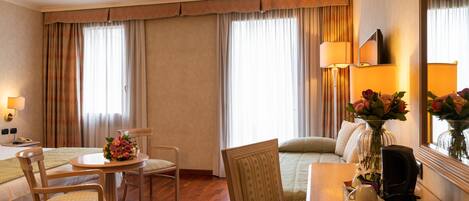 A hotel room with a bed, a desk with a chair, a small table with a vase of flowers, and a lamp.