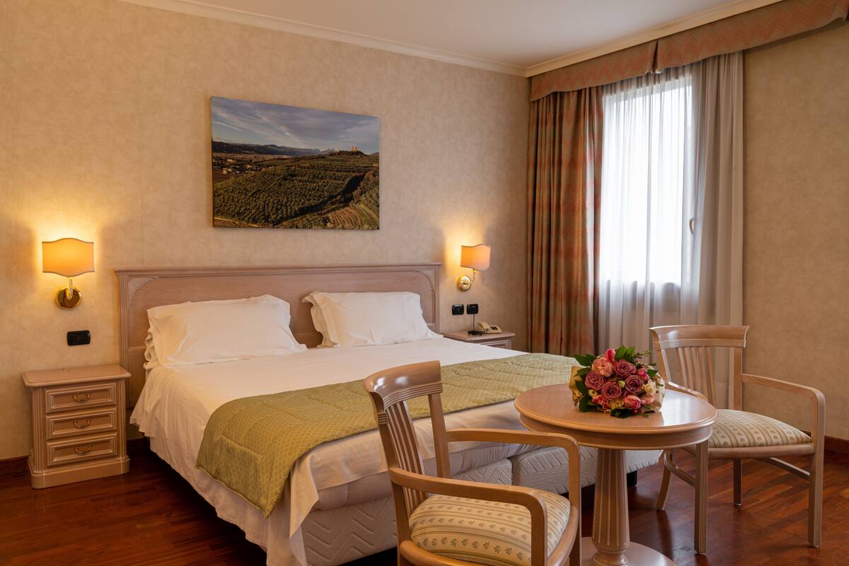 A hotel room with a large bed, two bedside tables with lamps, a small table with a floral arrangement, two chairs, and a painting on the wall.