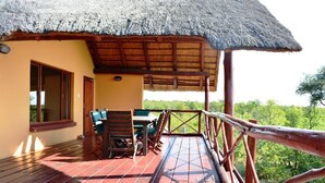 A wooden deck with a thatched roof structure, a table and chairs, and a view of the surrounding greenery.