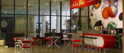 Una barra de jugos con un interior moderno, que incluye taburetes rojos y blancos, un amplio ventanal y un mural de frutas.