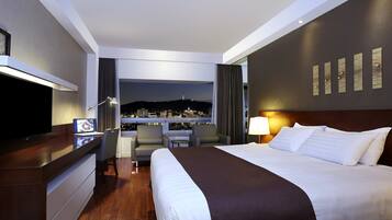 A modern hotel room with a large bed, a desk with a computer, a TV, and a view of a cityscape at night.