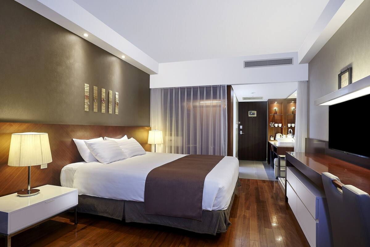 A modern hotel room with a large bed, bedside lamps, a flat-screen TV, a desk with a chair, and a bathroom visible through a glass door.