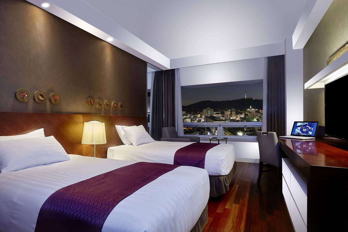 A hotel room with two beds, a desk, a chair, a TV, and a city view.