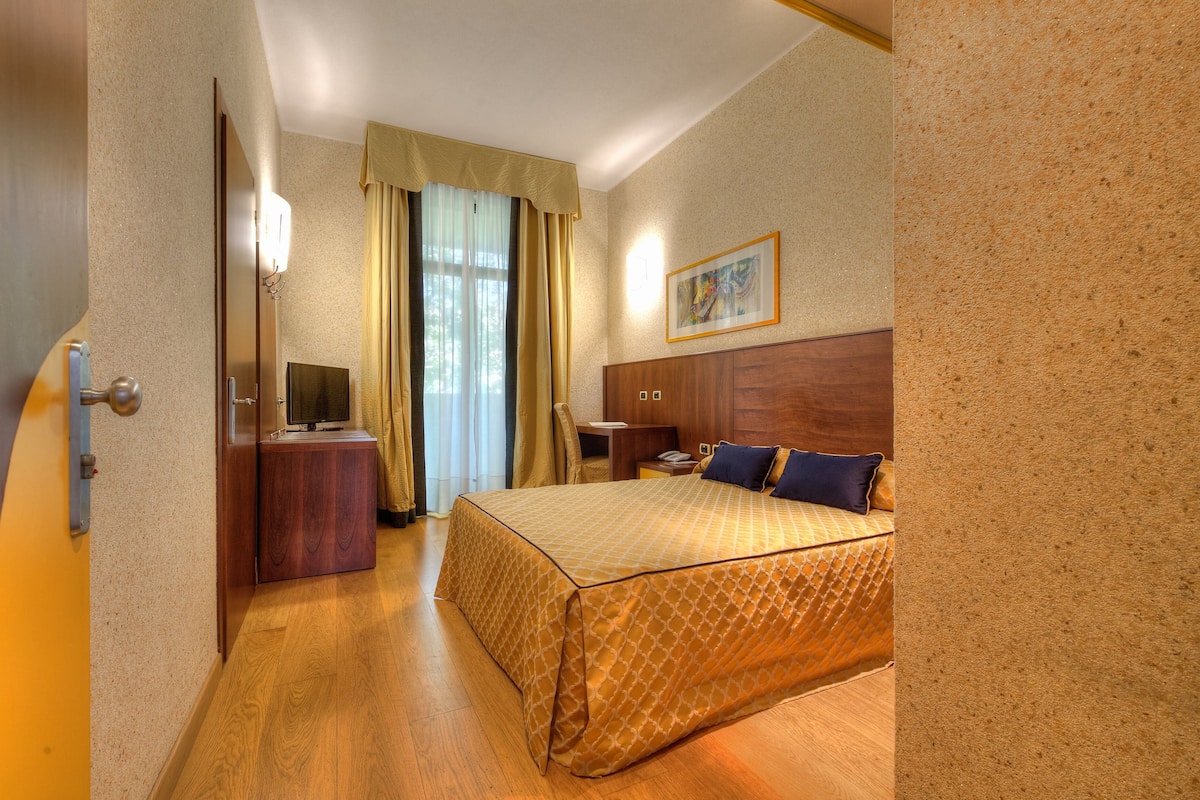 A hotel room with a bed, a desk with a television, a nightstand, and a painting on the wall.
