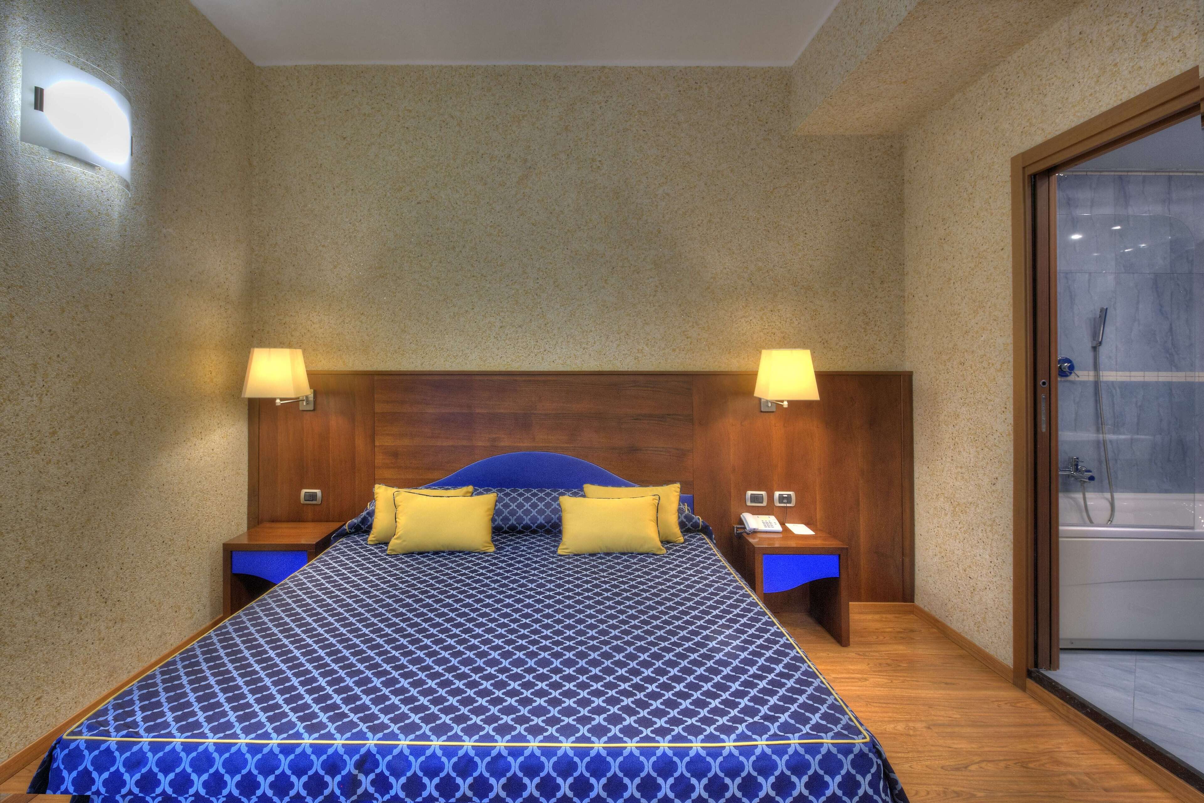 A hotel room with a large bed, two bedside tables, a wooden headboard, and a glass shower door.