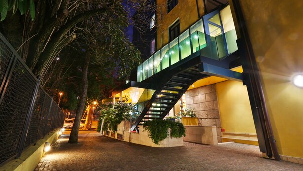 A nighttime view of a building with a glass bridge connecting two floors, surrounded by trees and a brick pathway. - Central Park Hotel Modena (Modena)