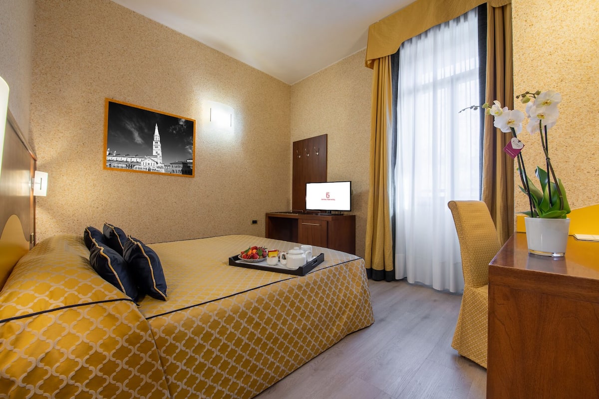 A hotel room with a bed, a desk, a chair, a TV, and a window with curtains.