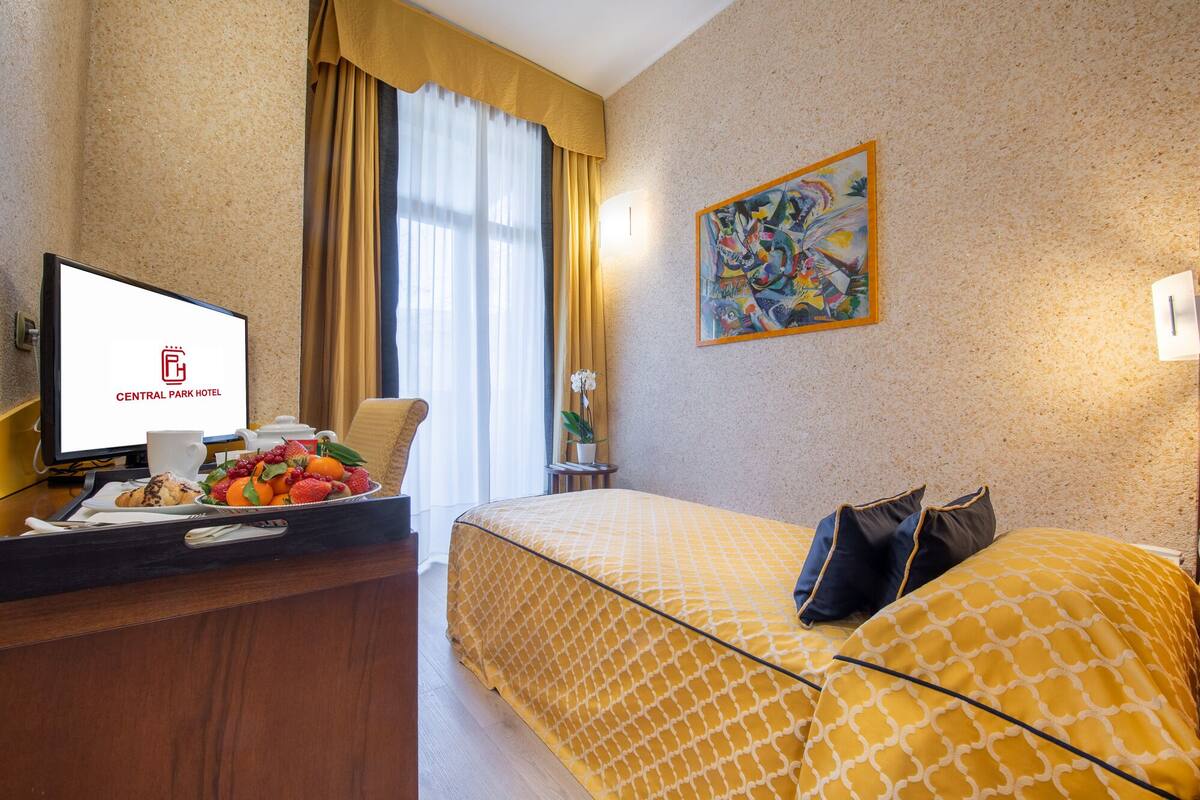 A hotel room with a bed, a desk with a computer, a painting on the wall, and a window with curtains.