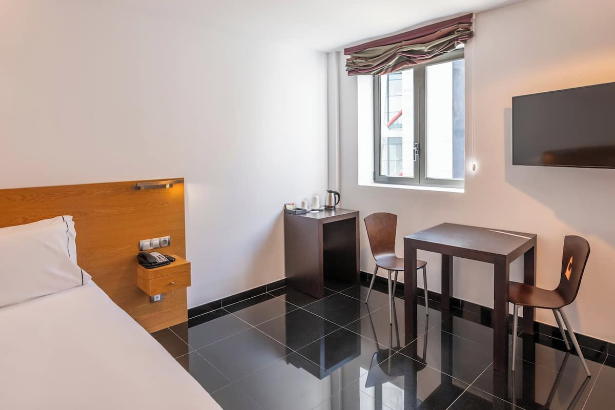 A hotel room with a bed, a desk with a chair, a small table, a telephone, and a window with curtains.