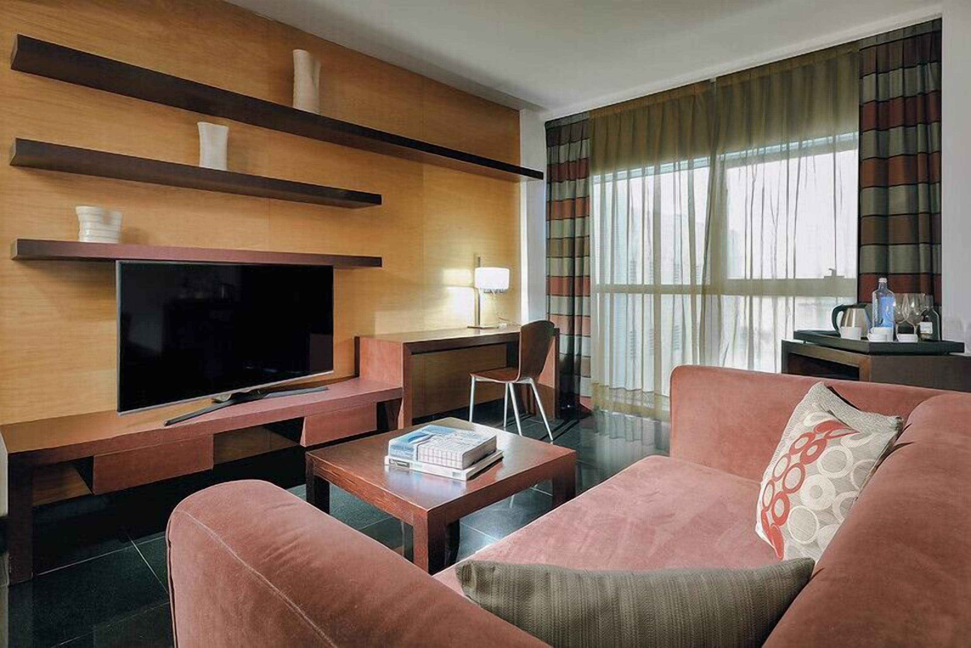 A modern living room with a flat-screen TV, wooden shelves, a sofa, a coffee table, and a desk with a lamp.