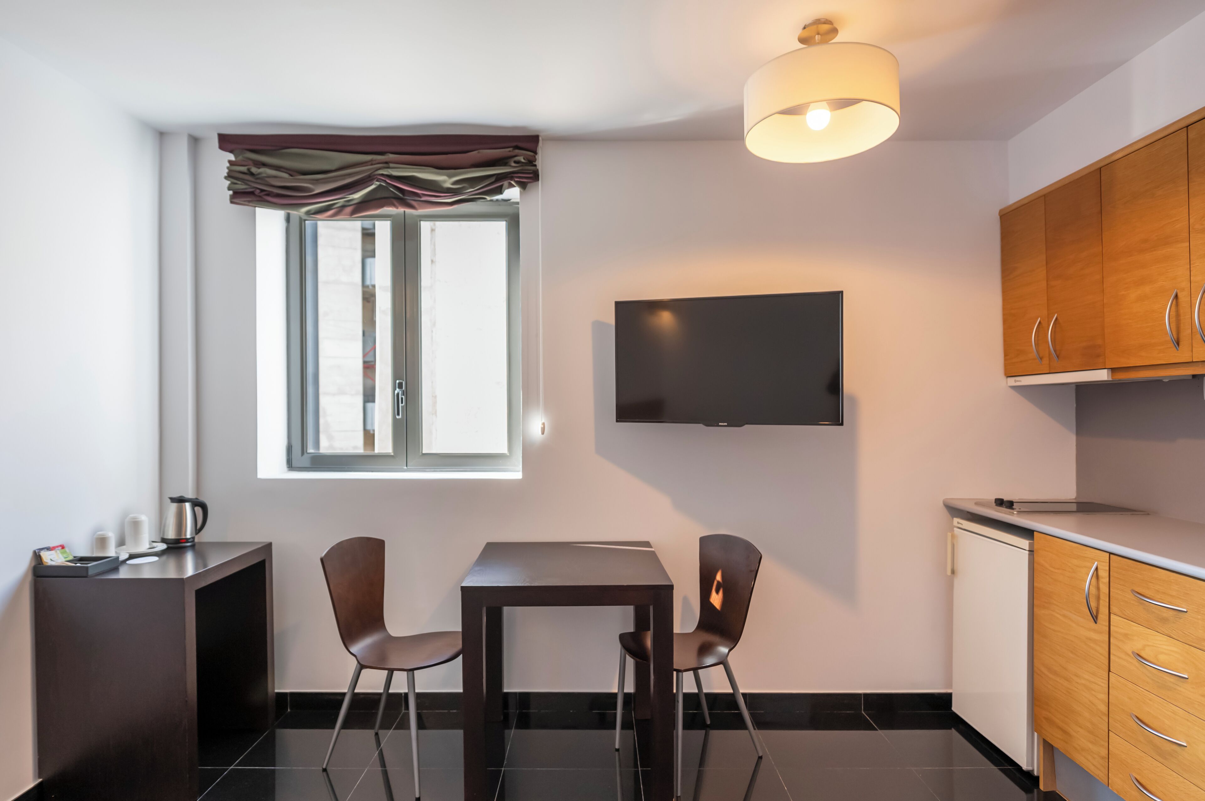 Una kitchenette moderna con un televisor de pantalla plana, dos sillas, una mesita y una ventana con cortinas.