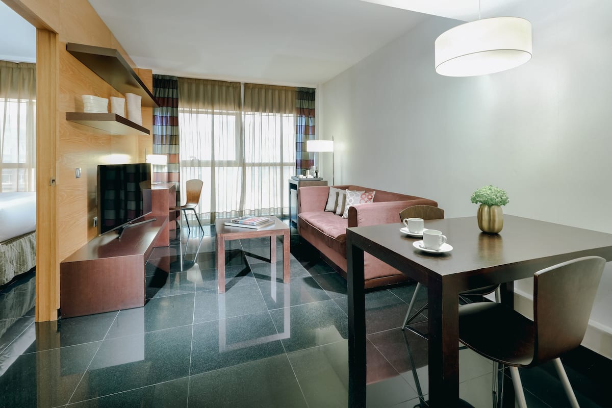 A modern hotel room with a sofa, a dining table with chairs, a TV, and a lamp.