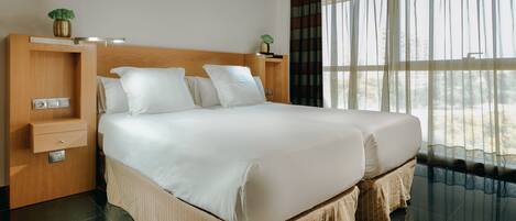A double bed with a wooden headboard, a bedside table with a plant, and a large window with curtains.