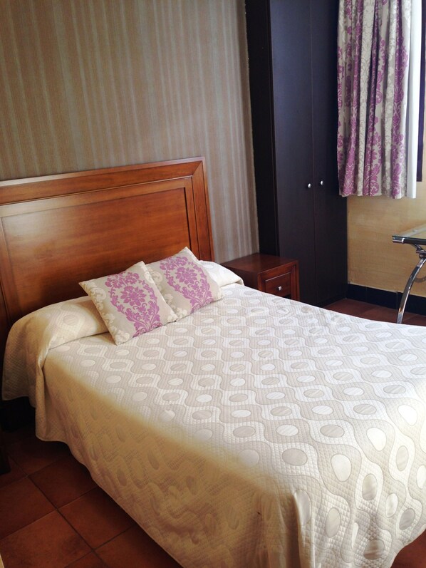 Una cama bien hecha con una colcha estampada y dos almohadas, un cabecero de madera, una mesita de noche y una ventana con cortinas.