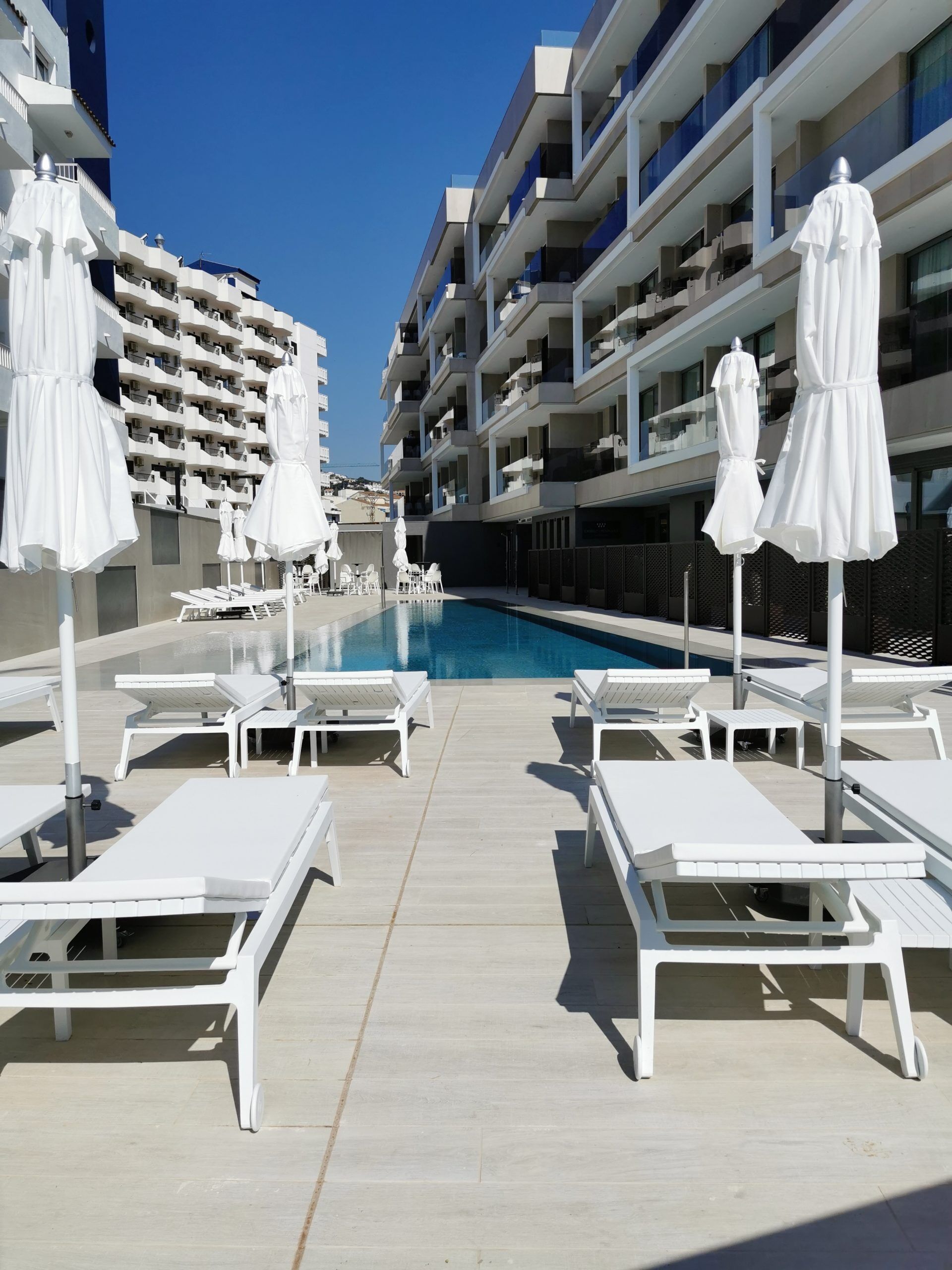 Un área junto a la piscina con sillas de descanso blancas y sombrillas, una piscina azul cristalina y edificios modernos de varios pisos al fondo.
