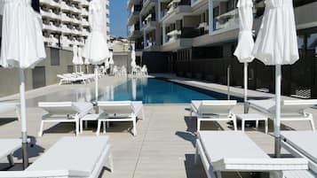 Un área junto a la piscina con sillas de descanso blancas y sombrillas, una piscina azul cristalina y edificios modernos de varios pisos al fondo.