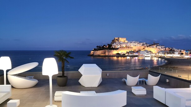 A rooftop terrace with modern white furniture, a palm tree, and a view of a historic fortified town by the sea. - Hotel & Spa Castillo de Peñíscola (Peniscola)