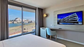 Een hotelkamer met een groot bed, een bureau, een stoel, een televisie waarop een gebouw wordt getoond, en uitzicht op een strand en stadsbeeld.