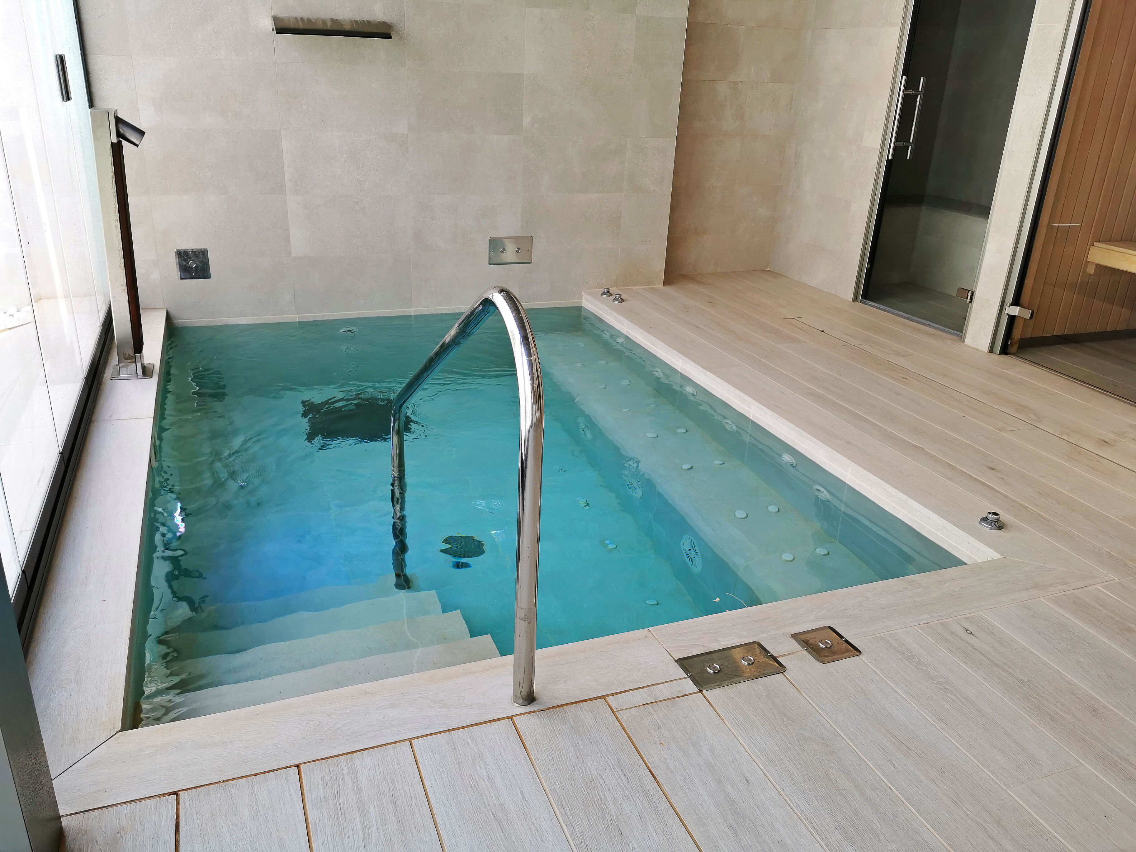 Una piscina con una escalera metálica, paredes de azulejos y una zona de terraza de madera.