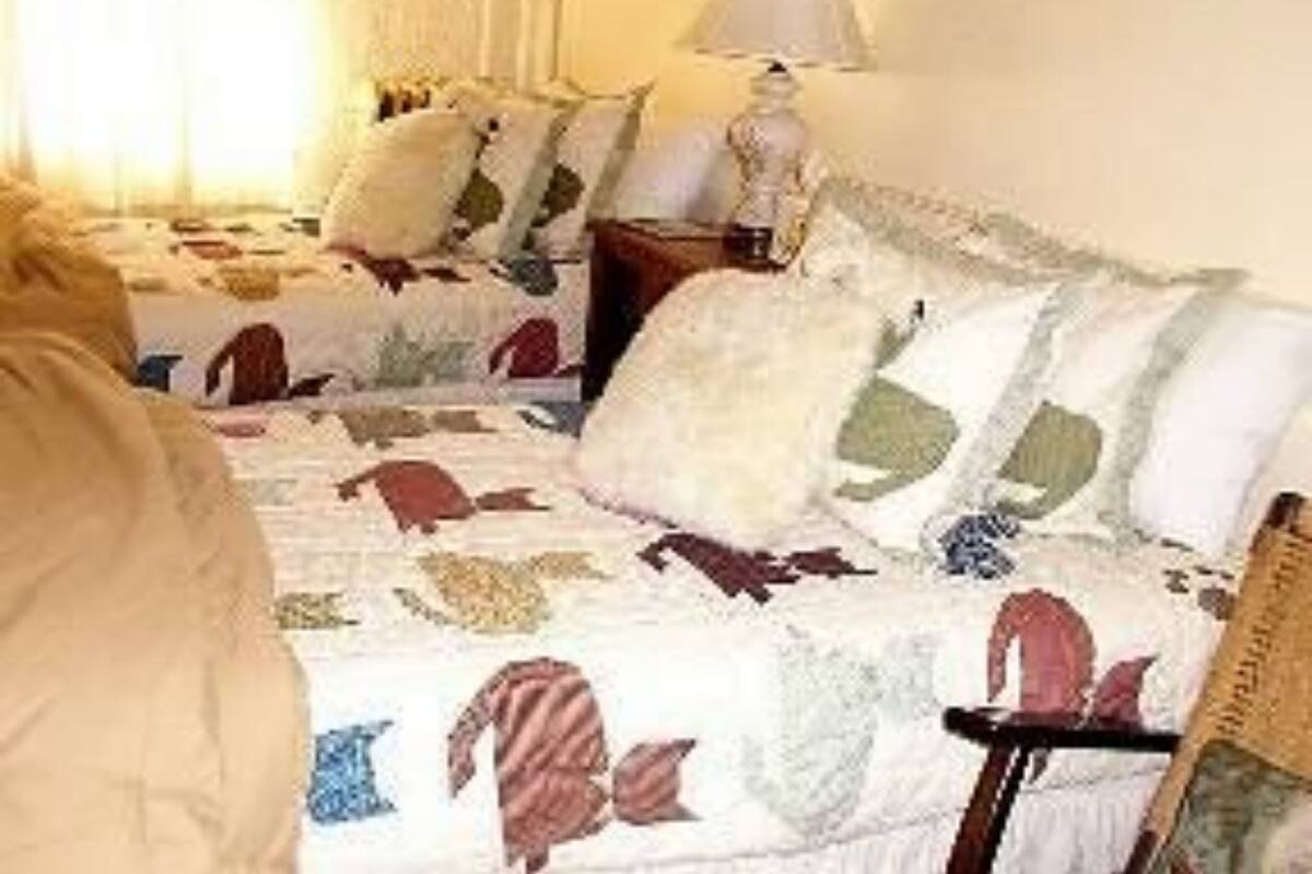 A bedroom with two beds, each with a patterned quilt. There is a bedside table with a lamp and a chair.