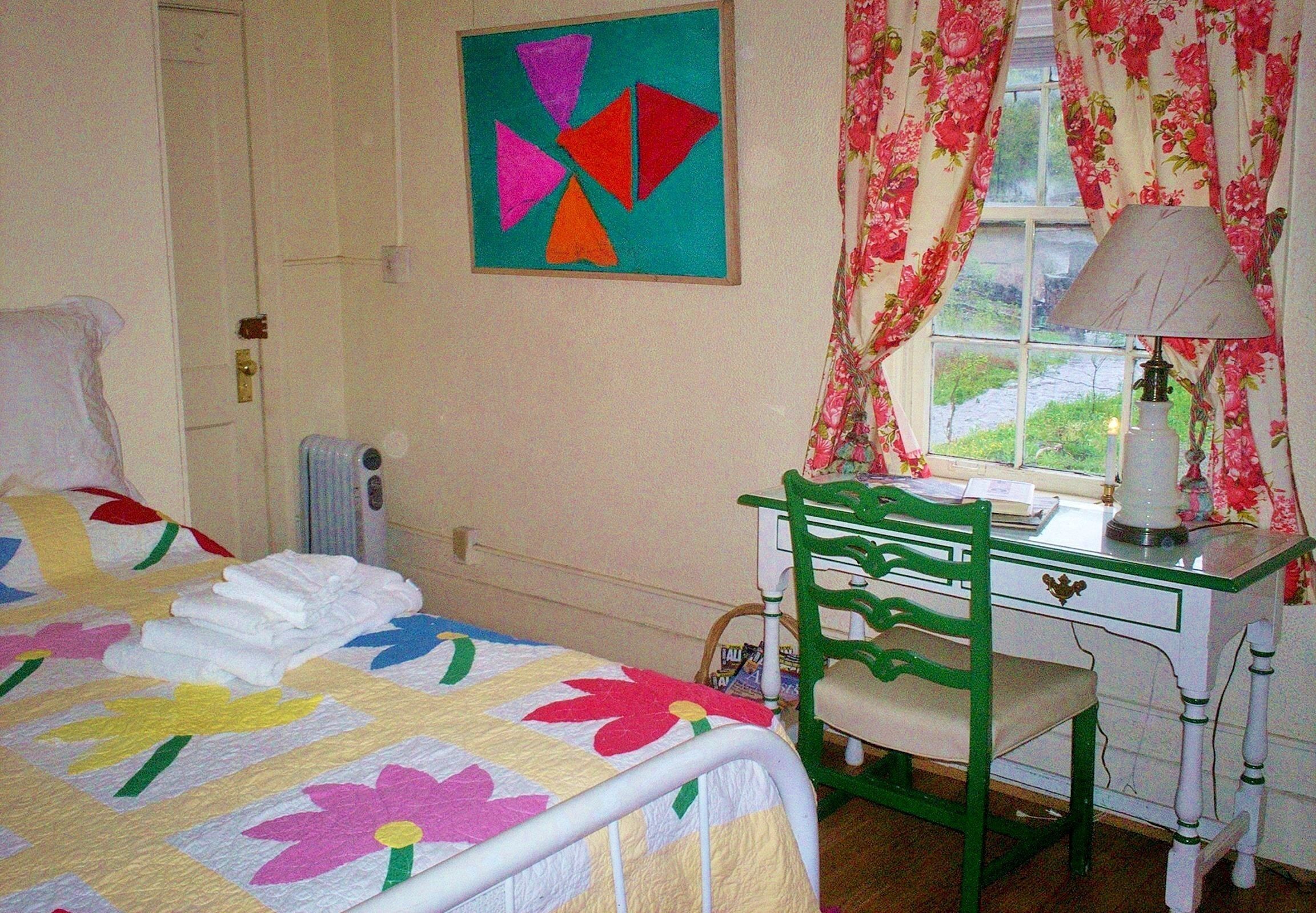 A bedroom with a bed, a desk with a chair, a window with curtains, and a painting on the wall.