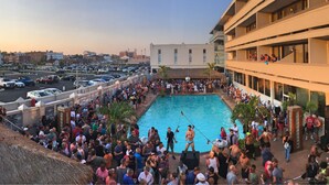 A crowded outdoor swimming pool area with a large crowd of people, a parking lot, and a building with multiple floors and balconies.