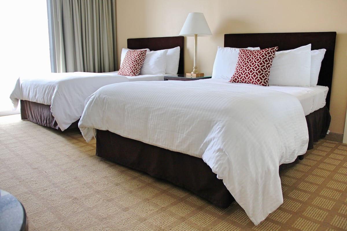 A hotel room with two beds, a nightstand, a lamp, and a view of the outdoors.