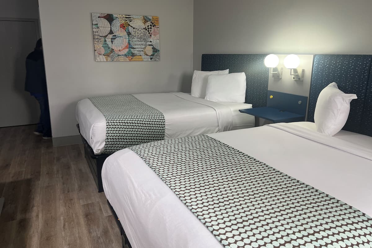 A hotel room with two beds, a nightstand, a wall-mounted lamp, and a painting.
