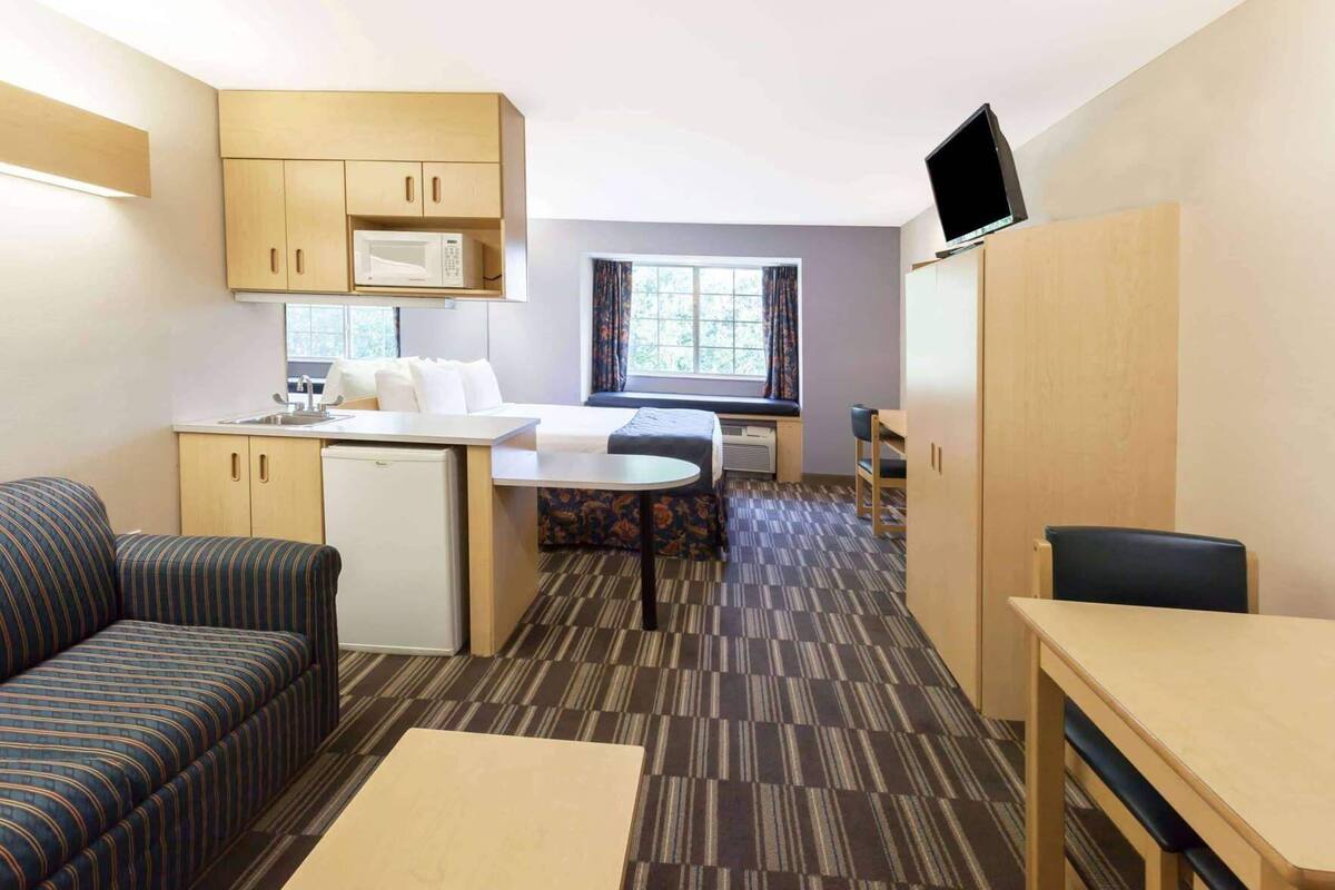 A compact hotel room with a kitchenette, a bed, a sofa, a dining table, and a TV.