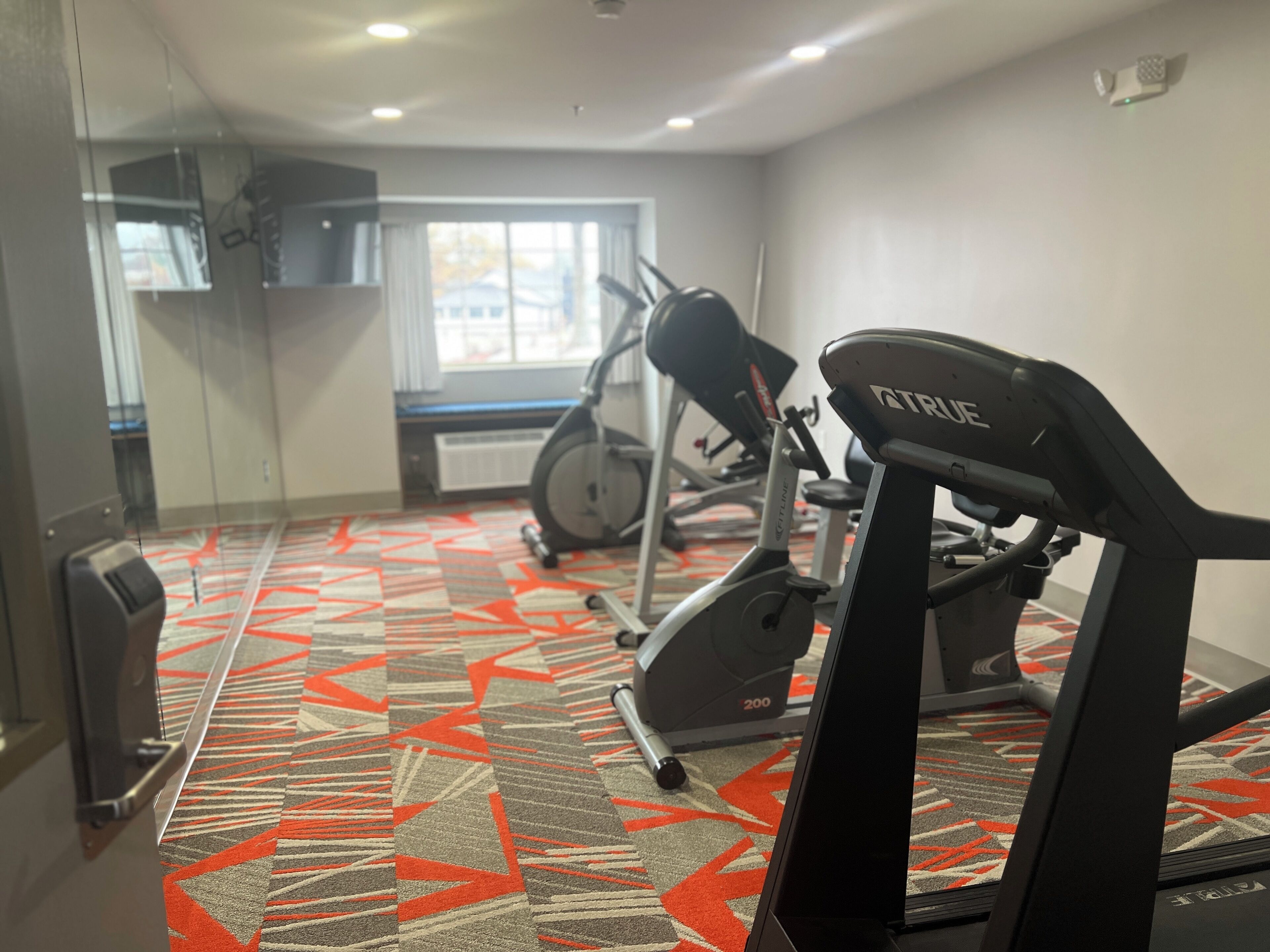 A gym with exercise equipment, a patterned carpet, and a window with a view of buildings.