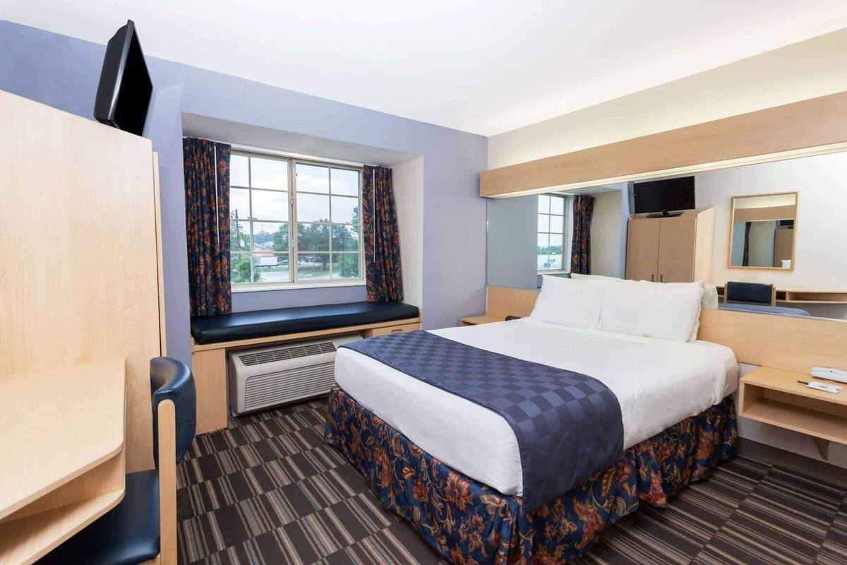 A hotel room with a bed, a desk, a chair, a window with curtains, a television, and a mirror.