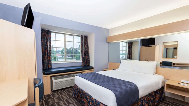 A hotel room with a bed, a desk, a chair, a window with curtains, a mirror, and a television on the wall.