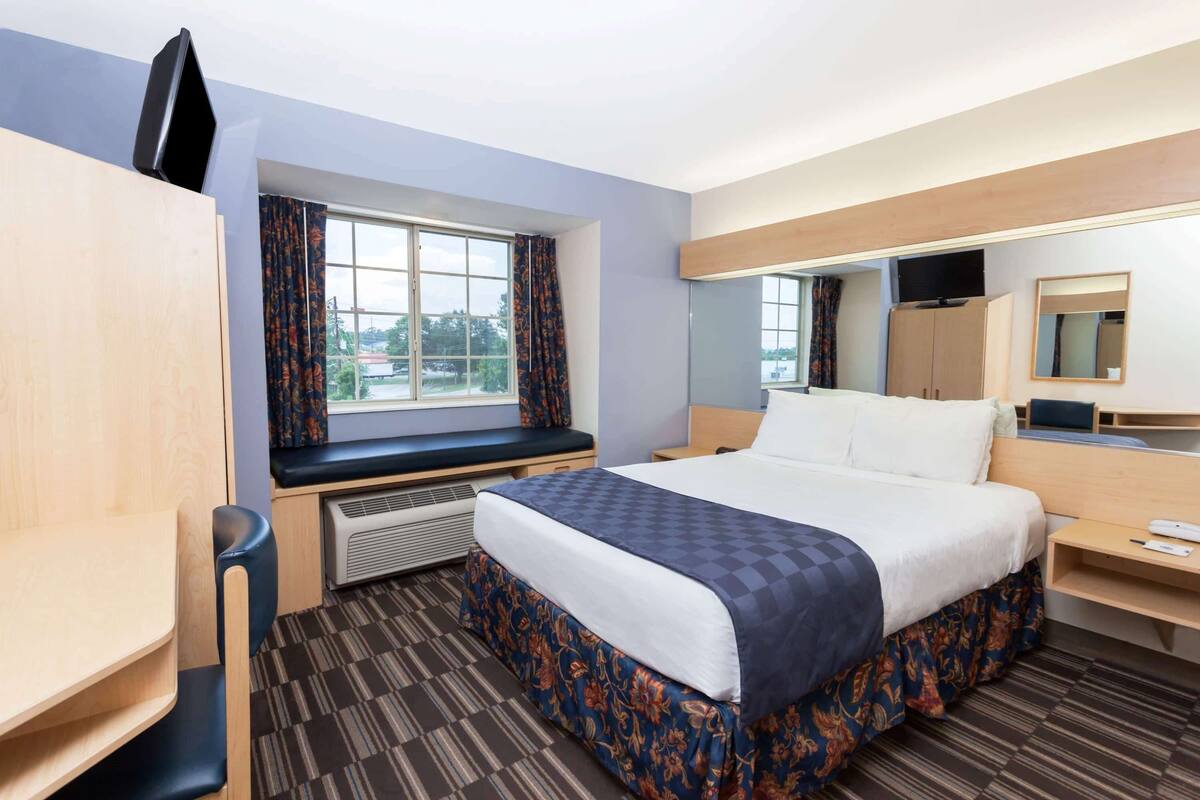 A hotel room with a bed, a desk, a chair, a window with curtains, a mirror, and a television on the wall.