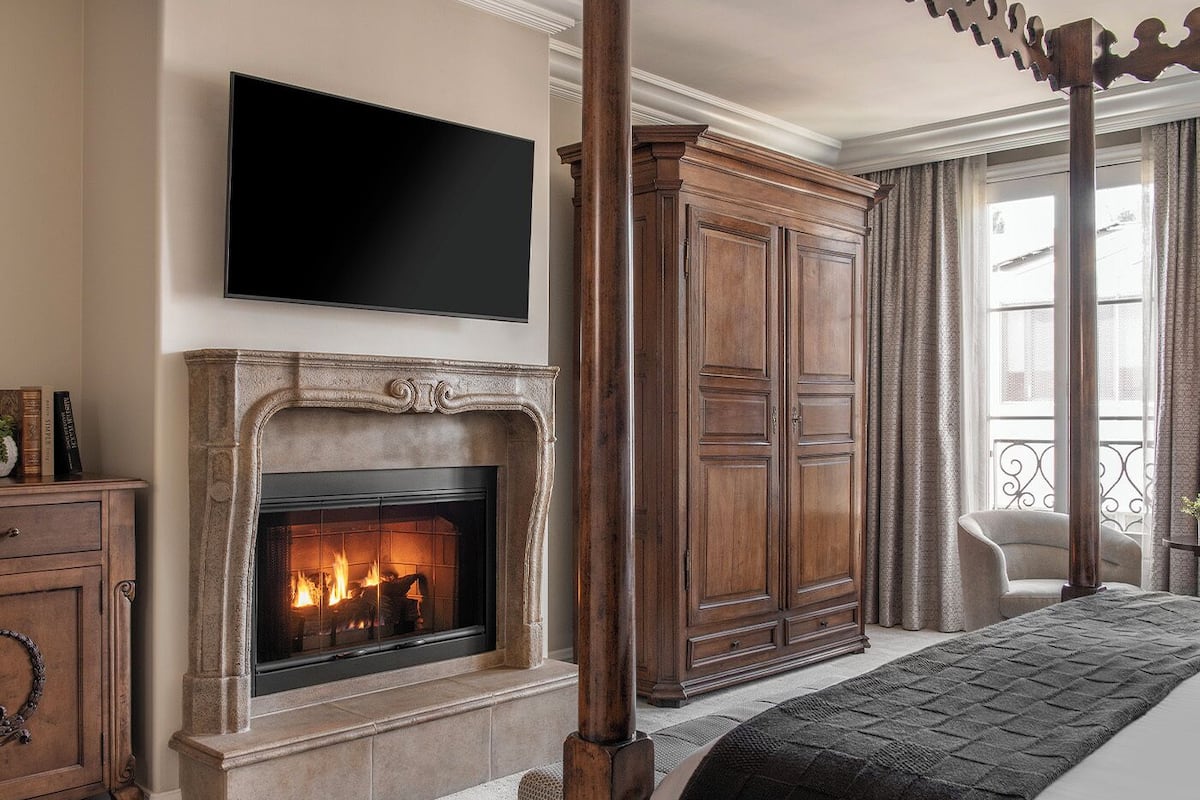 Ein Schlafzimmer mit Kamin, einem darüber montierten Fernseher, einem großen Holzschrank und einem Bett mit dunkler Decke.