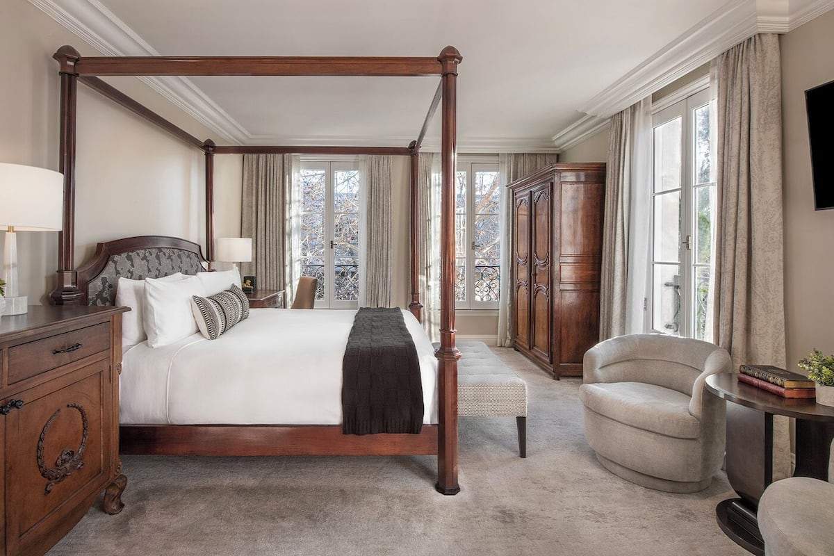 Ein Bett mit Himmel, dessen dunkles Holzgestell erkennbar ist, ein Nachttisch mit Lampe, ein Sessel, ein Kleiderschrank und ein Blick ins Freie durch ein großes Fenster.