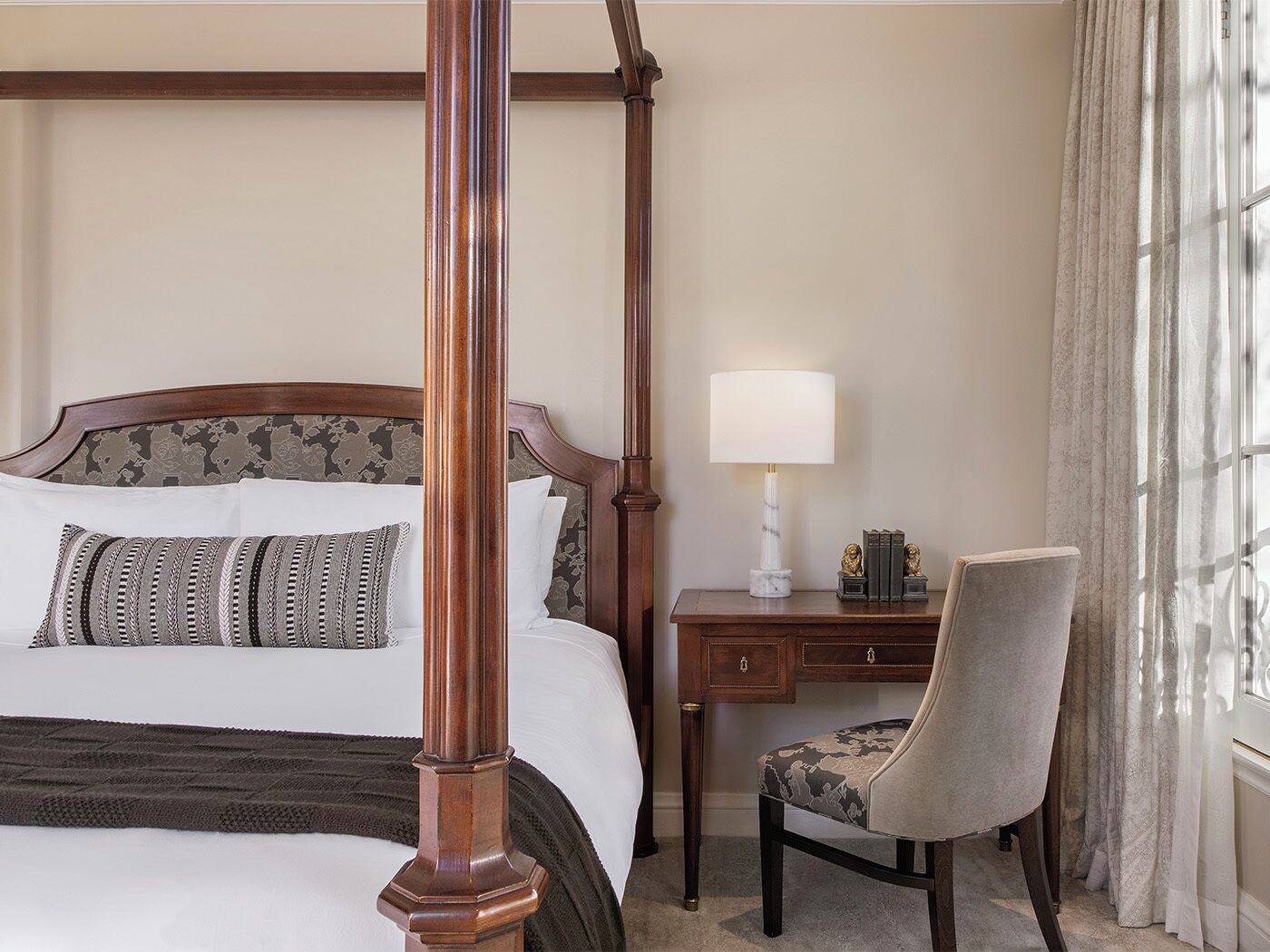 A four-poster bed with a headboard, a bedside table with a lamp, a chair, and a window with curtains.