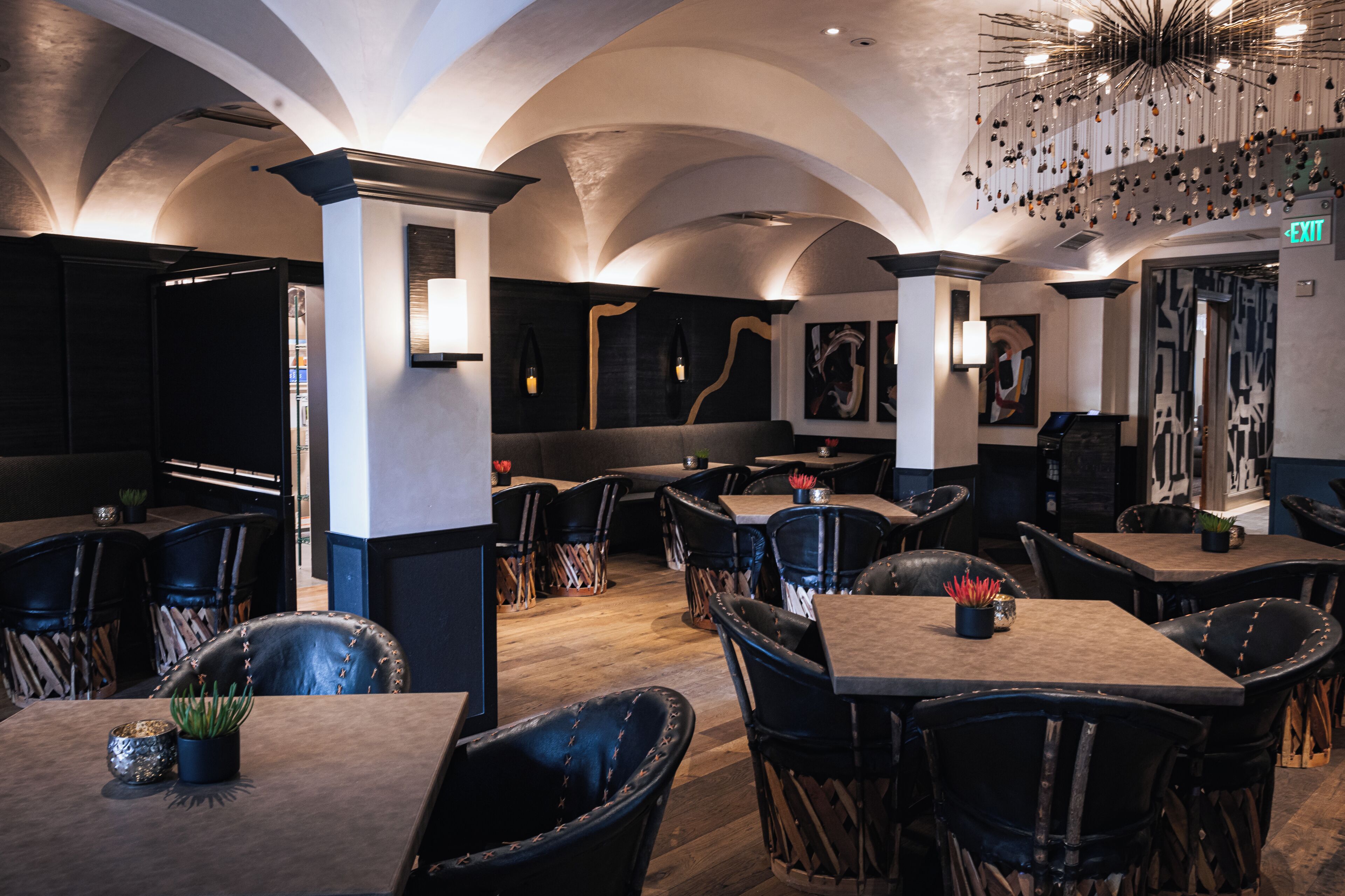 Un interno ristorante con soffitti a volta alta, sedie in pelle nera e tavoli con piante in vaso.