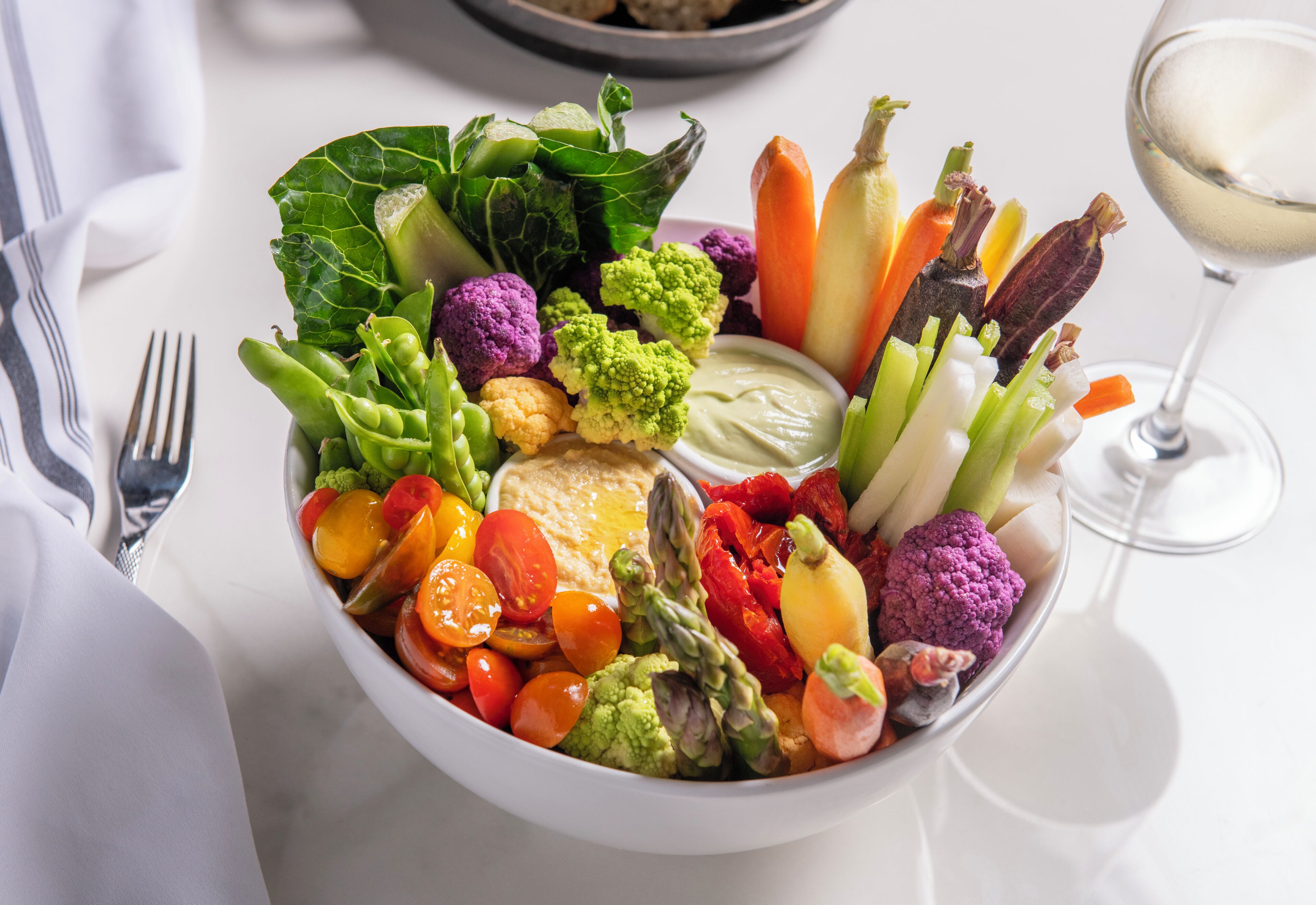 A bowl of assorted vegetables and a glass of white wine on a table.