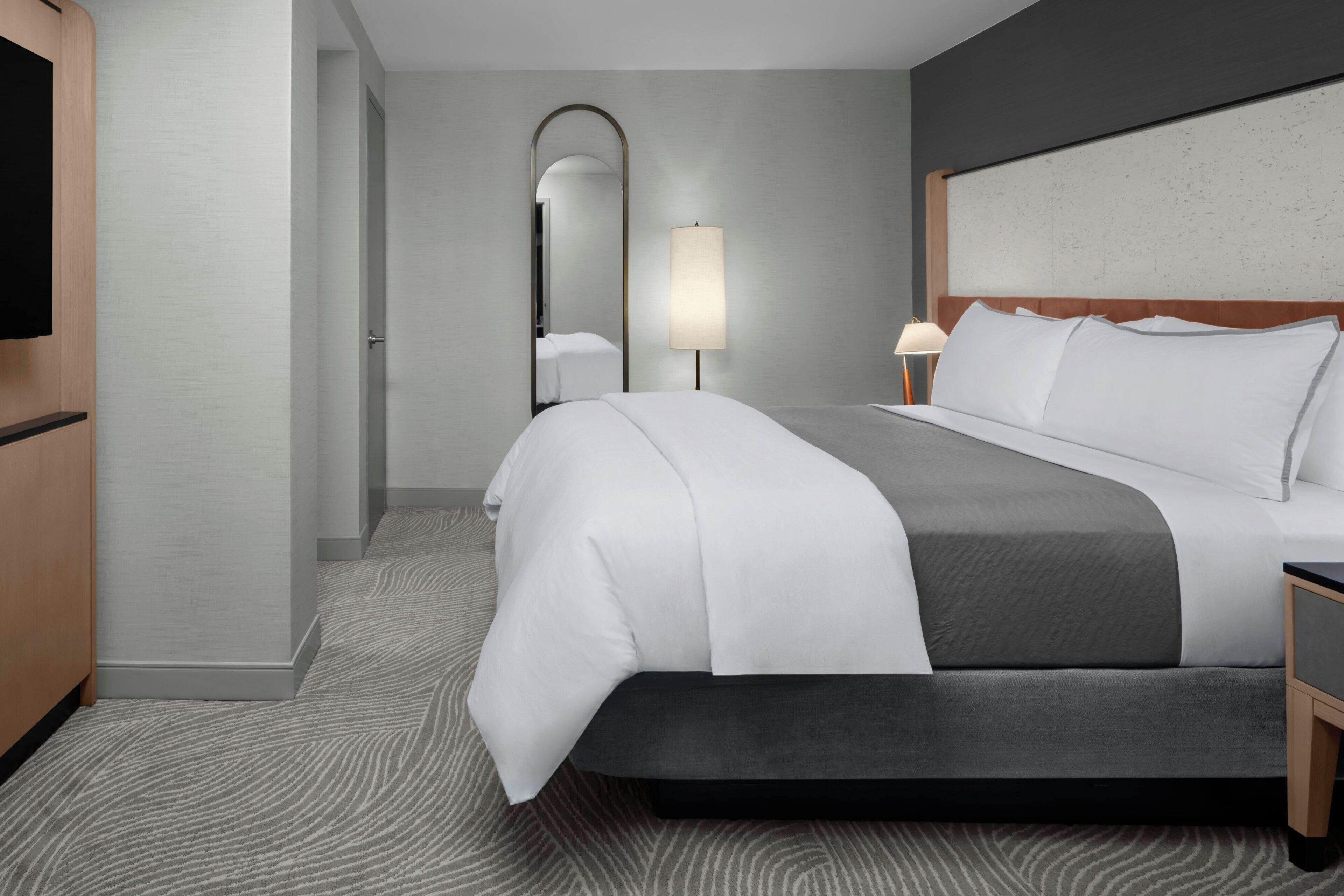 A hotel room with a bed, a TV, a mirror, and a lamp.