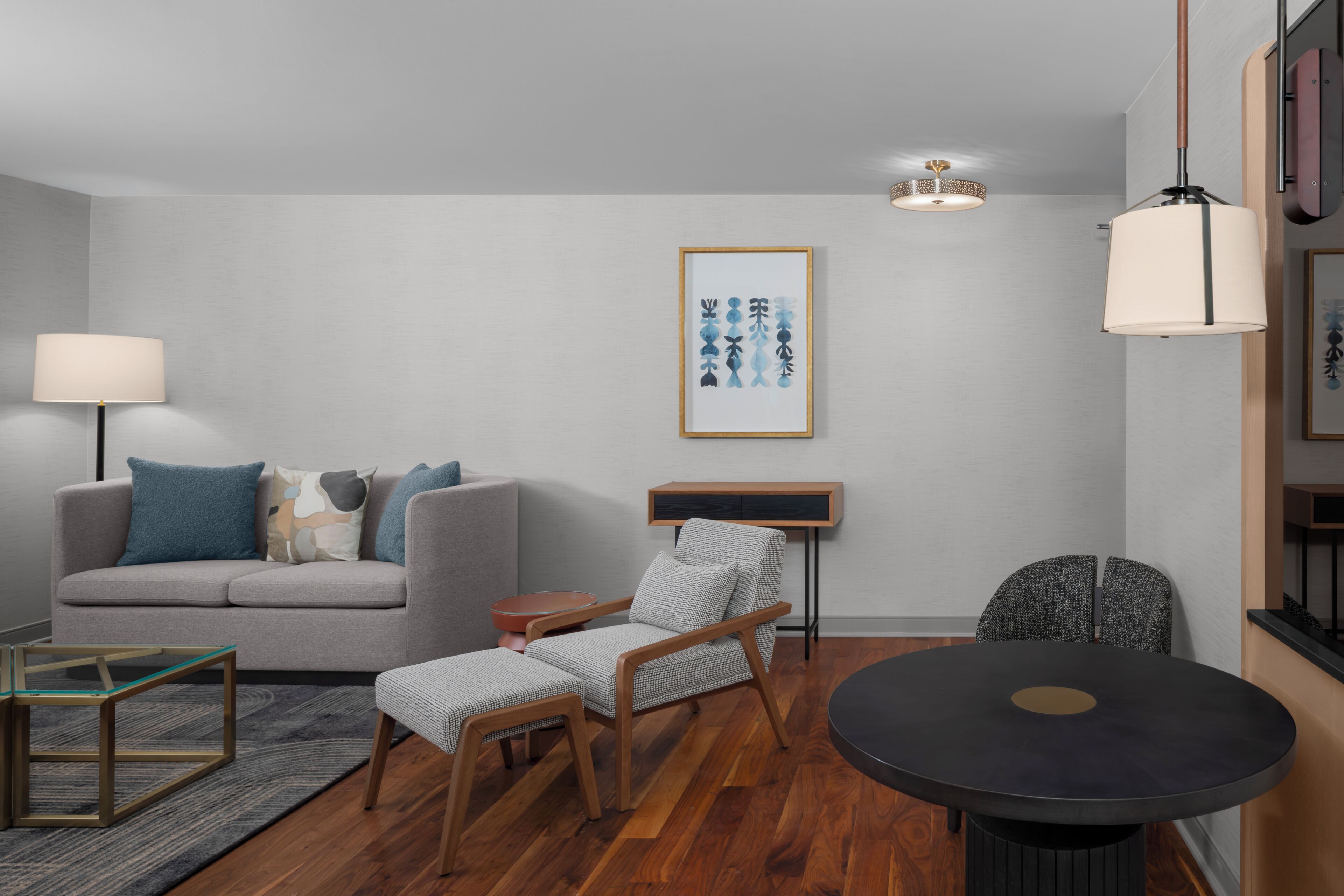 A modern living room with a grey sofa, a small round table, a wooden chair, a floor lamp, and a framed artwork on the wall.