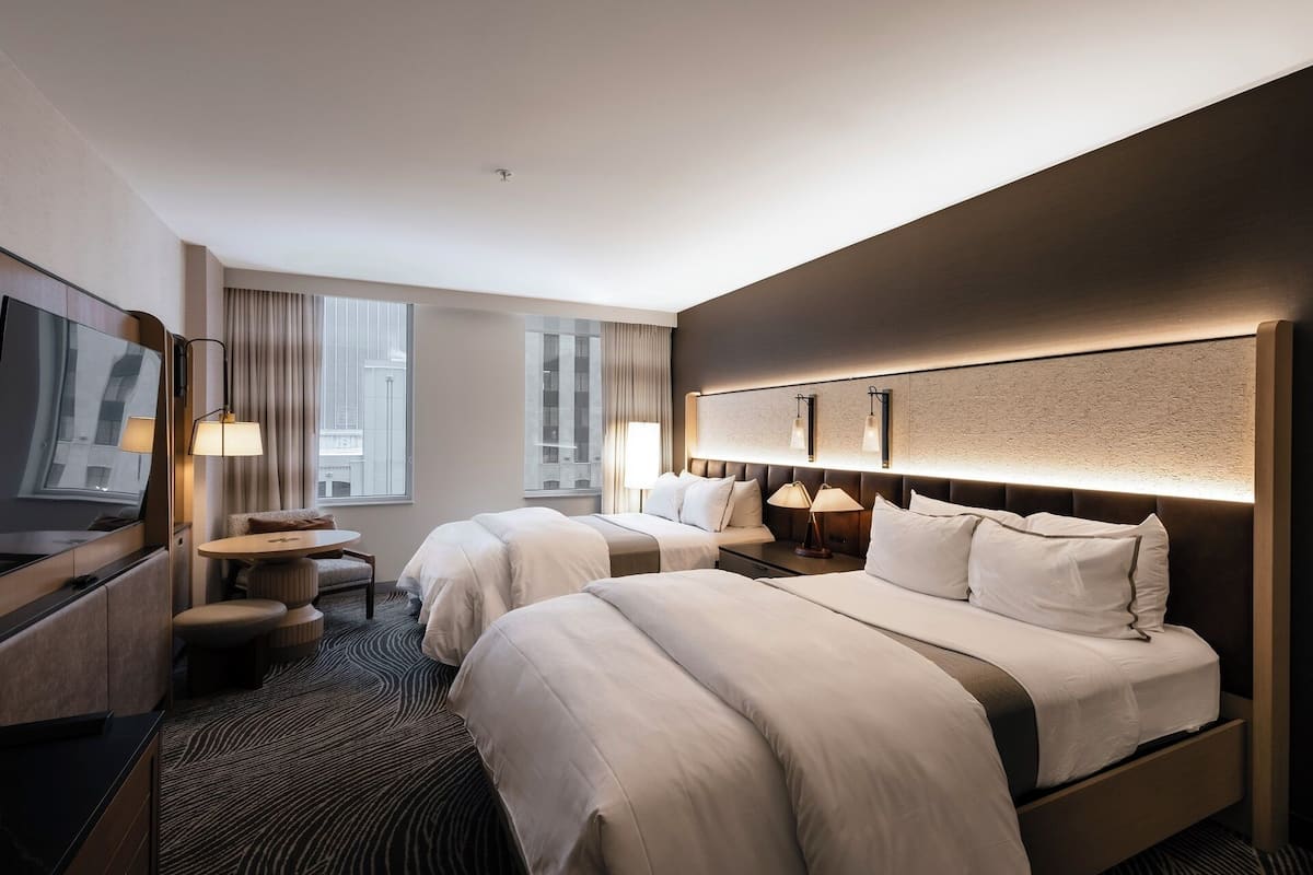 A hotel room with two beds, a desk, a chair, a TV, and a large window.