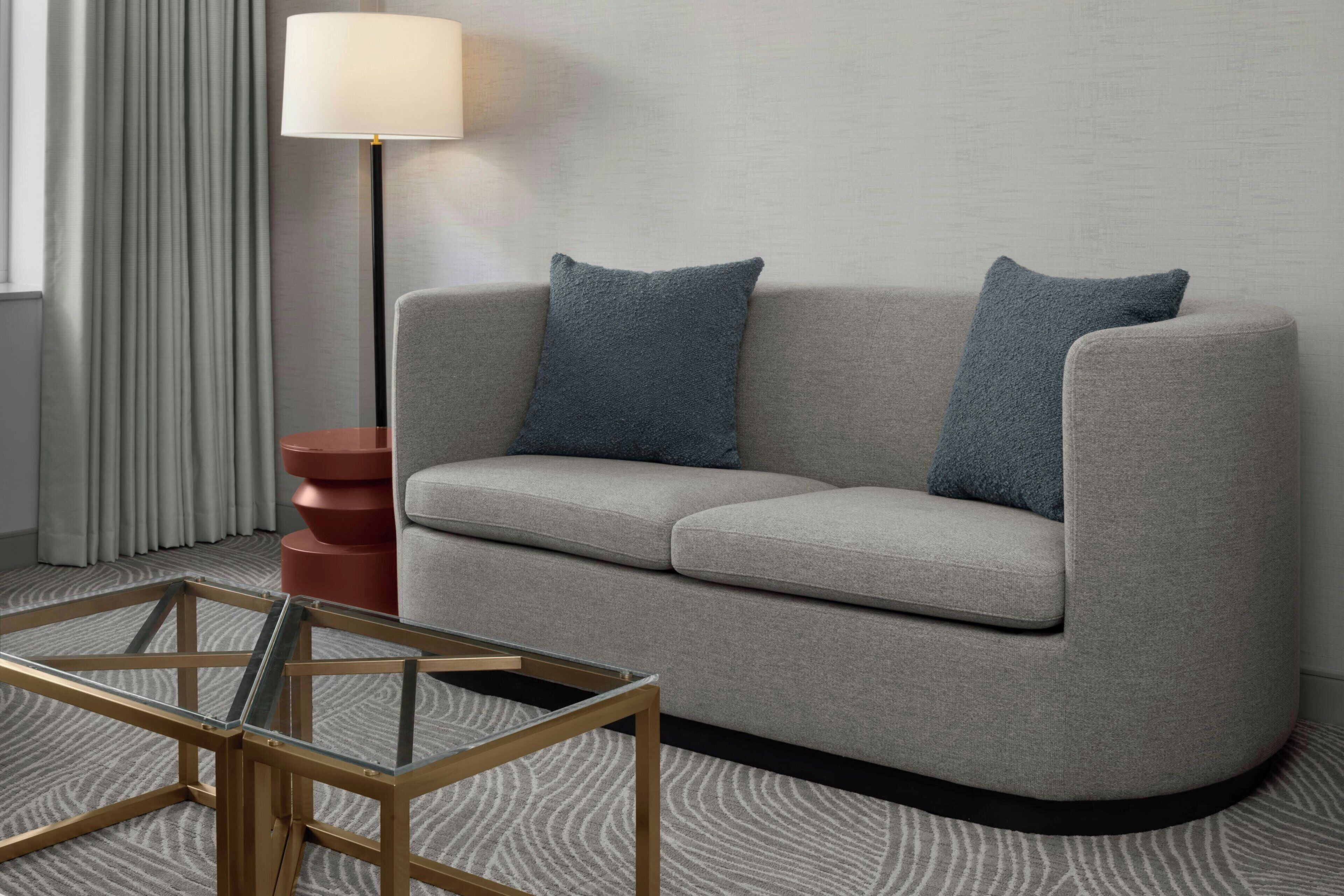 Una sala de estar moderna con un sofá gris, dos cojines azules, una mesa de centro de vidrio, una lámpara de pie y una alfombra estampada.