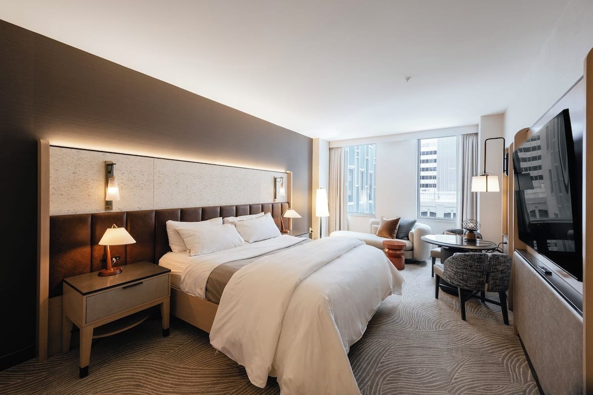 A hotel room with a large bed, bedside tables, a desk, a chair, a TV, and a window with curtains.