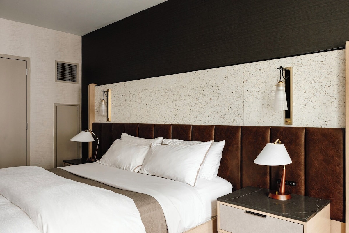 A hotel room with a bed, bedside tables, a headboard, and a wall-mounted light fixture.