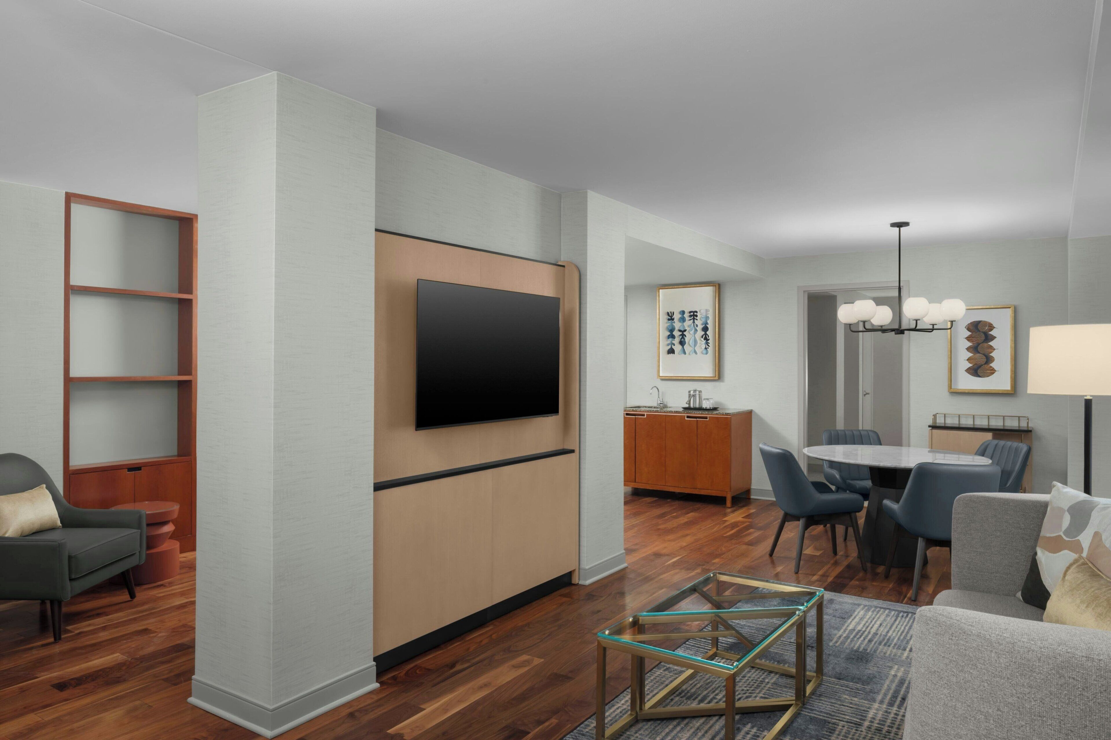 Uma sala de estar moderna com televisão de tela plana, uma área de jantar com mesa e cadeiras redondas, uma cozinha com armários de madeira e um corredor que dá para outro quarto.