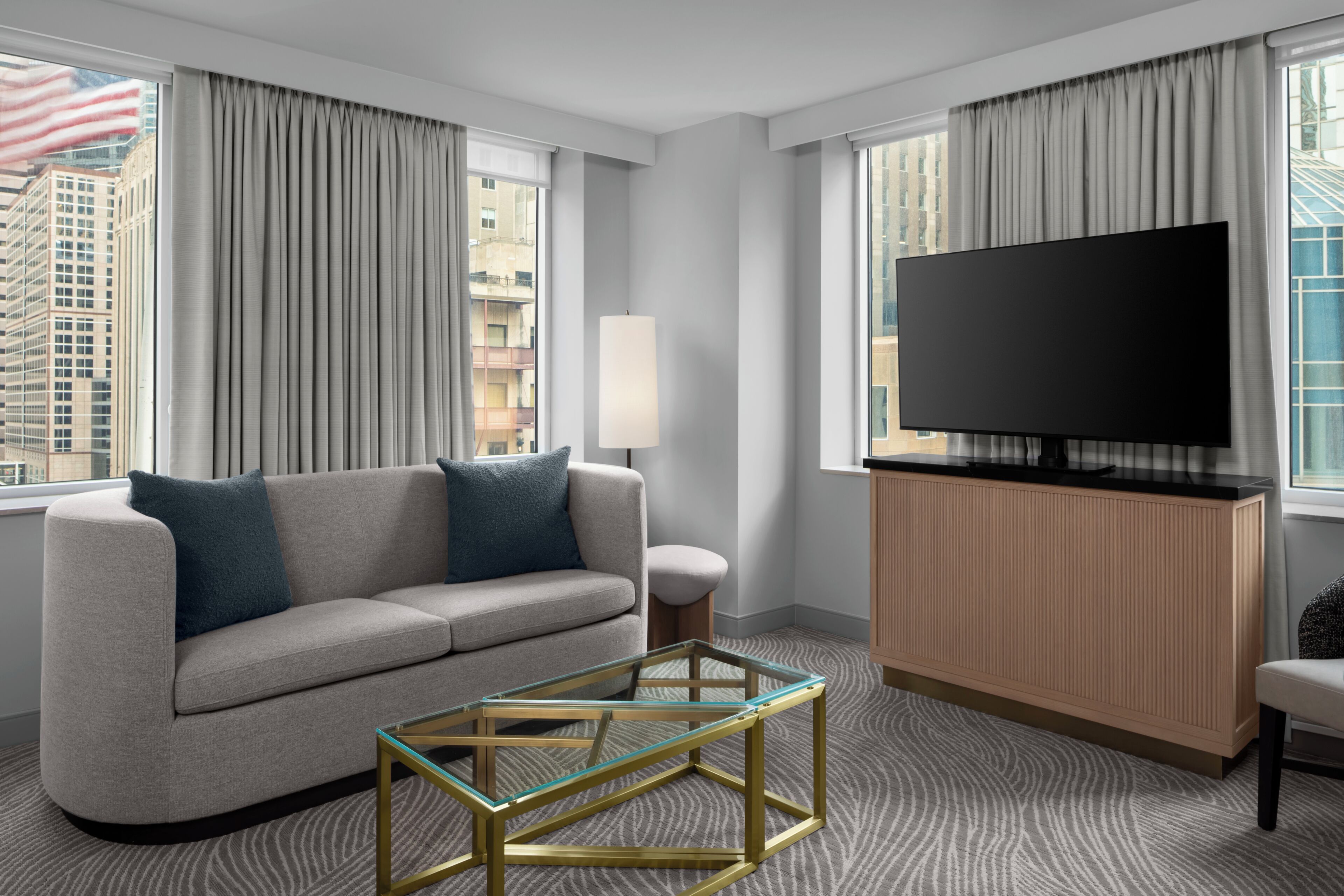 A modern hotel room with a sofa, a glass coffee table, a flat-screen TV, and large windows with curtains.